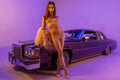 A woman posing in a purple bikini with a fringe detail, standing next to a vintage purple car under purple studio lighting.